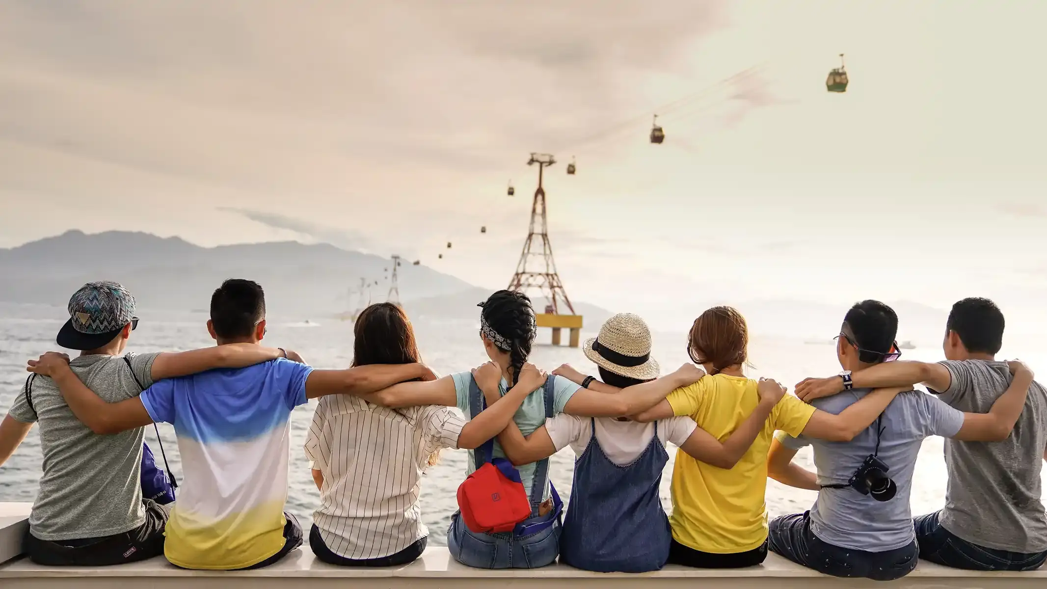 Friends sitting arm in arm on a sea wall, looking out at the water and a cable car