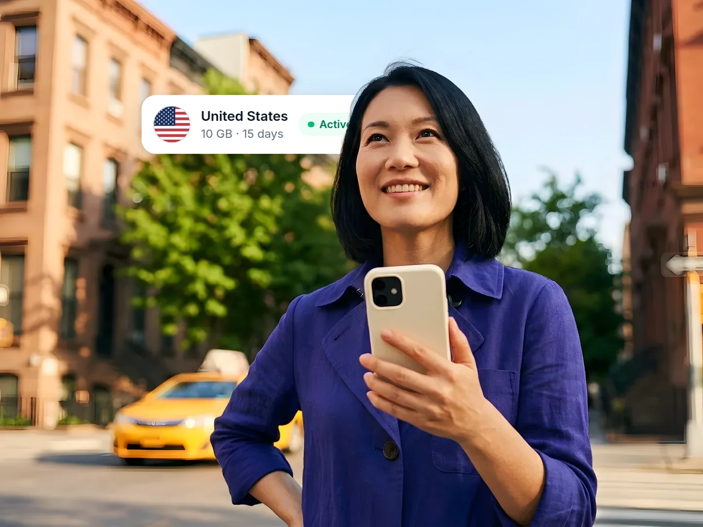 Woman in an indigo coat holding her phone on a sunny New York street, smiling up at the city, beside an active United States eSIM card